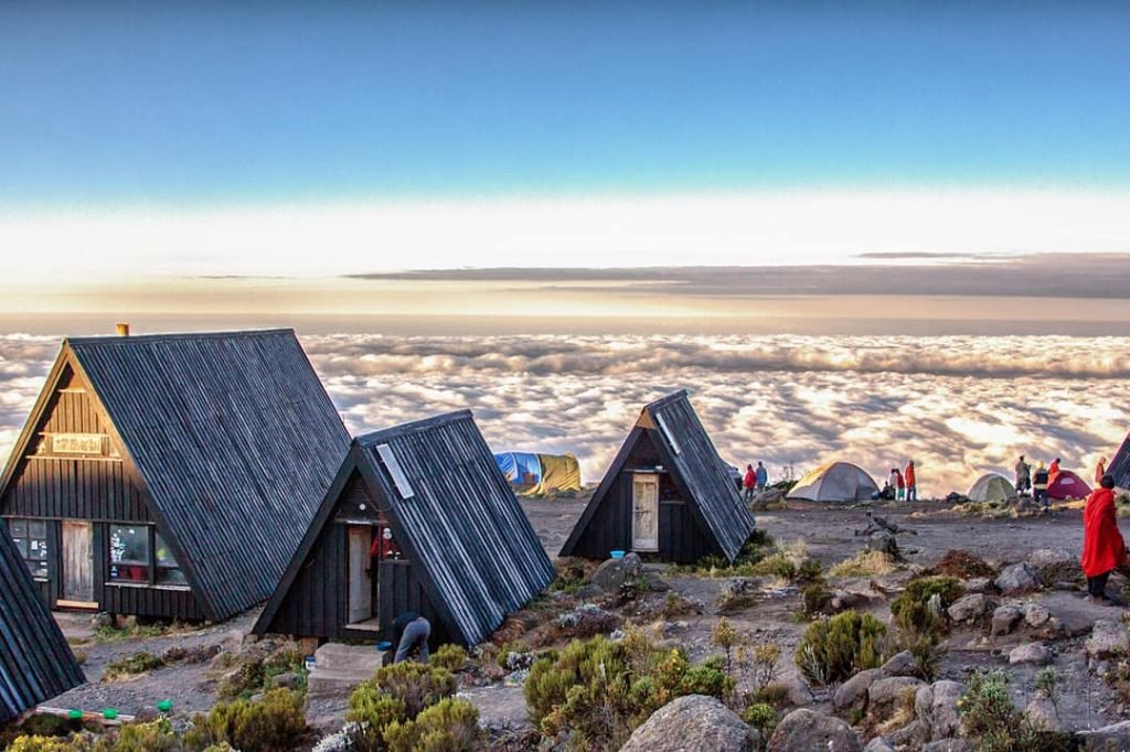 A row of Marangu route accommodation huts on Mount Kilimanjaro. The huts are made of woods and have thatched roofs. They are situated on a hillside overlooking a lush green valley.