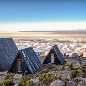 A row of Marangu route accommodation huts on Mount Kilimanjaro. The huts are made of woods and have thatched roofs. They are situated on a hillside overlooking a lush green valley.