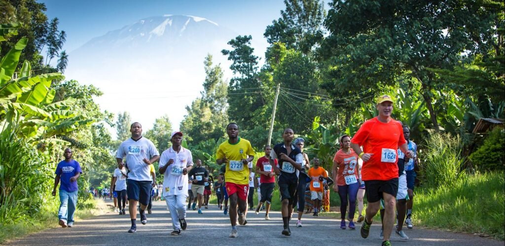 Runners participating in the Kili Marathon with Mount Kilimanjaro in the background. The image captures a vibrant and dynamic scene of the marathon event, showcasing participants in athletic gear, the majestic snow-capped peak of Kilimanjaro, and the scenic landscape of Tanzania. Ideal for highlighting adventure sports, marathon events, and travel experiences in Africa.